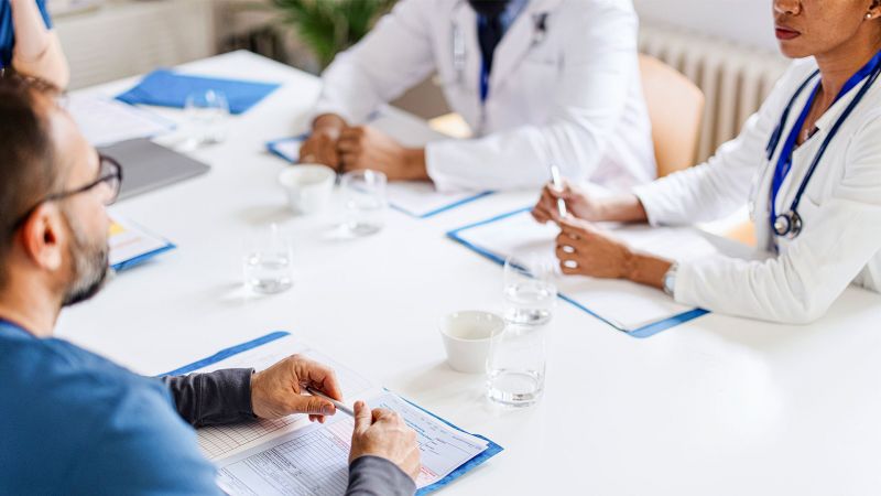 Photo; team of doctors and the monitor sit at a table and look through patient files.