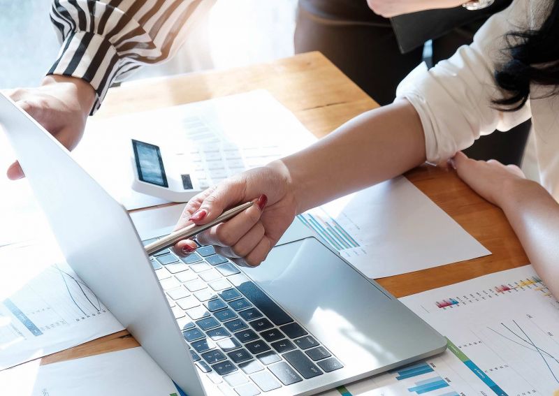 Photo; a person points to a laptop with a pen; on the table are reports with diagrams
