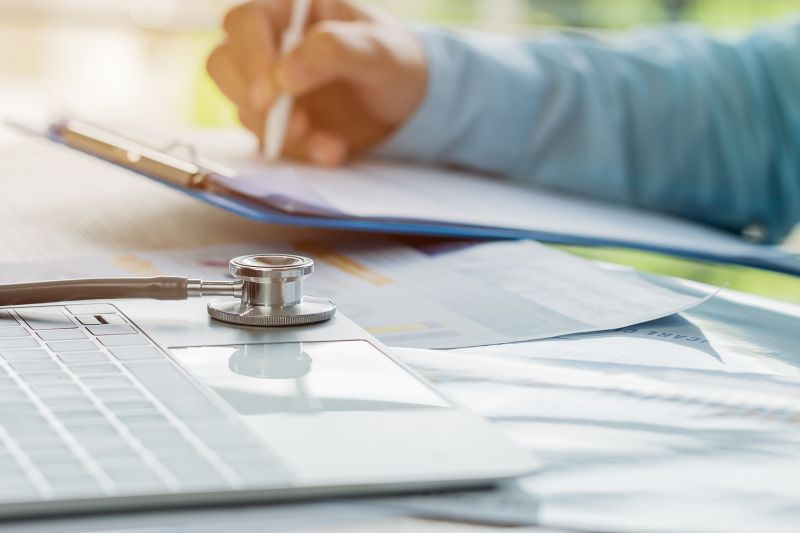 Photo; laptop with stethoscope on keyboard, behind it a hand holding a pen on a clipboard.